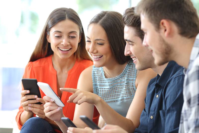 Happy group of friends checking smart phones sitting on a couch in the living room at home