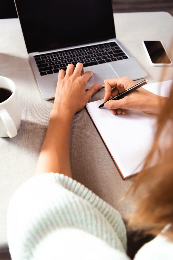 woman writing in notepad in cafe with coffee cup, smartphone and laptop